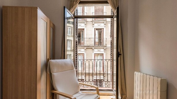Une pièce avec une armoire en bois, un fauteuil blanc rembourré, un radiateur et une vue sur un immeuble par une porte-fenêtre donnant sur un balcon.