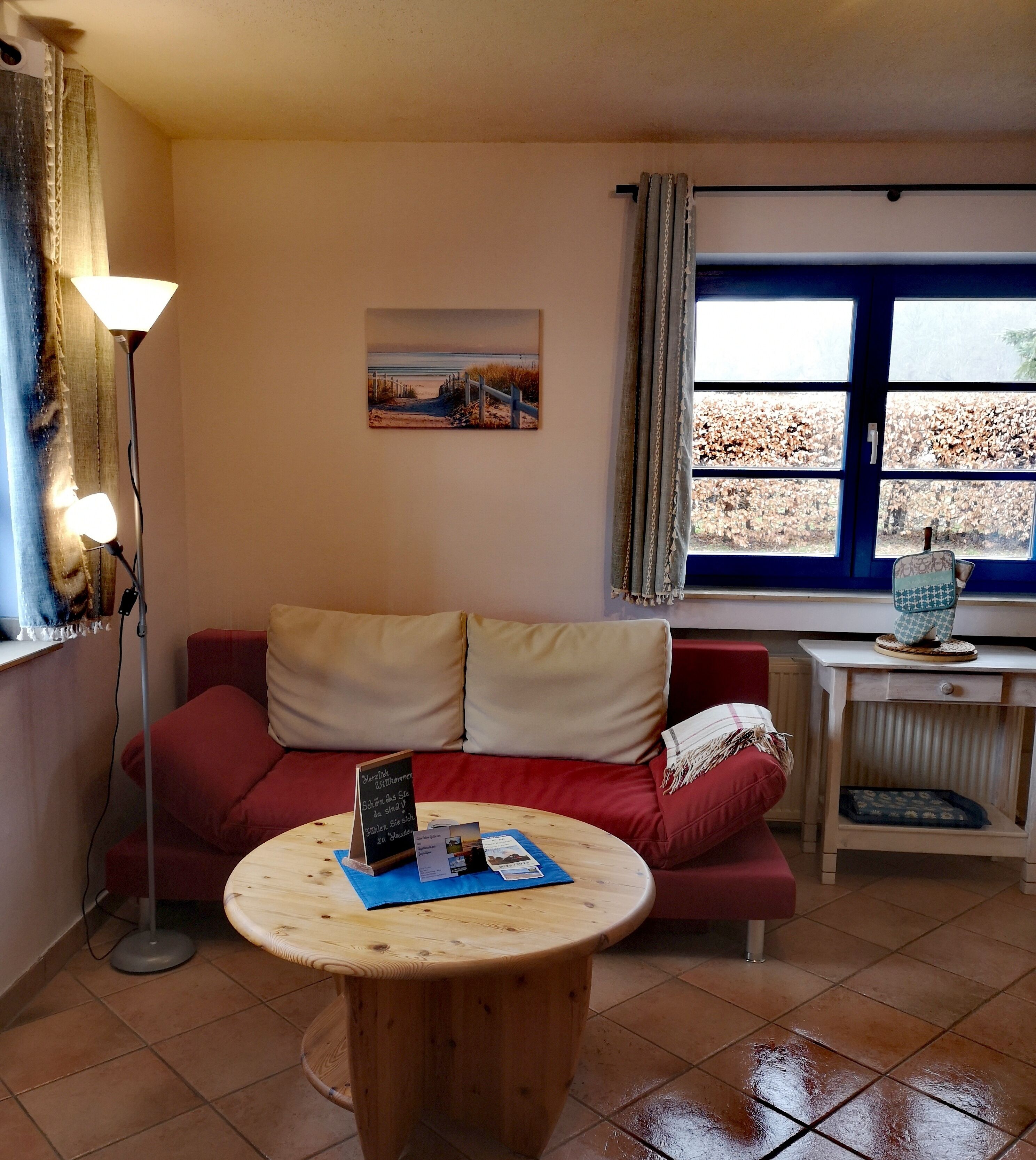 Ein gemütliches Wohnzimmer mit einem roten Sofa, einem runden Holztisch, einer Lampe, einem Bild und einem Fenster mit Blick auf Bäume.