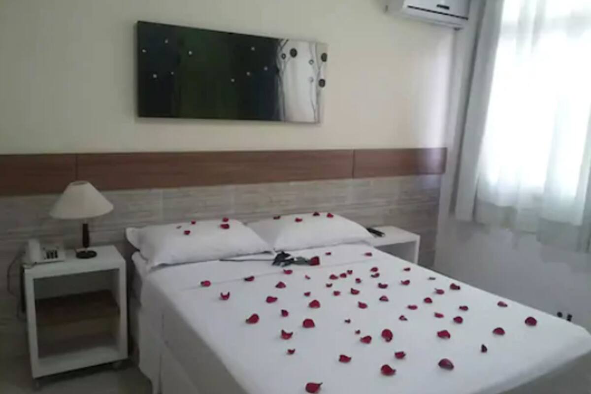 A hotel room with a bed, a nightstand, a lamp, an air conditioner, and a painting on the wall.