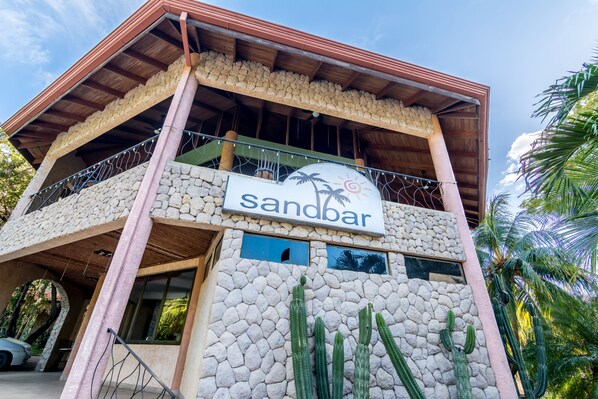 A two-story building with a sign reading "Sandbar." It features a stone facade, a balcony with a metal railing, and several cacti in front.