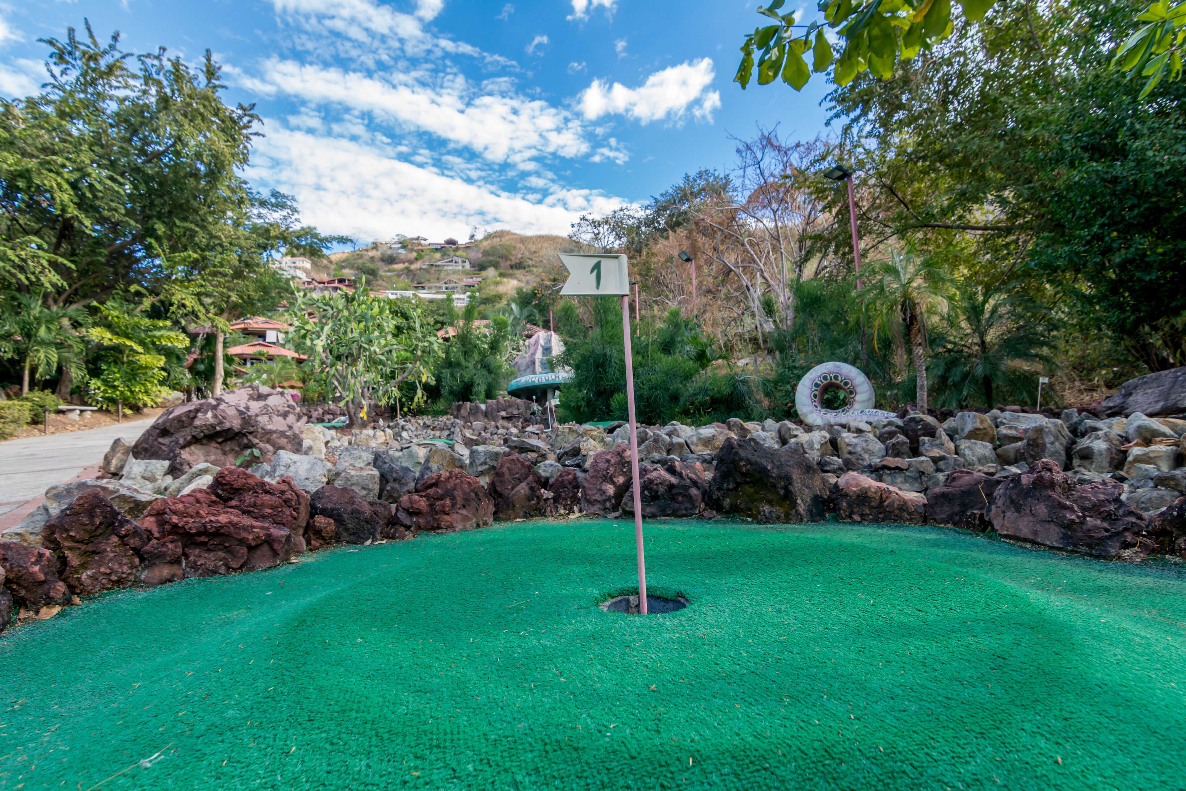 A miniature golf course with a green playing surface, a hole marked by a flag, and a stone wall in the background.