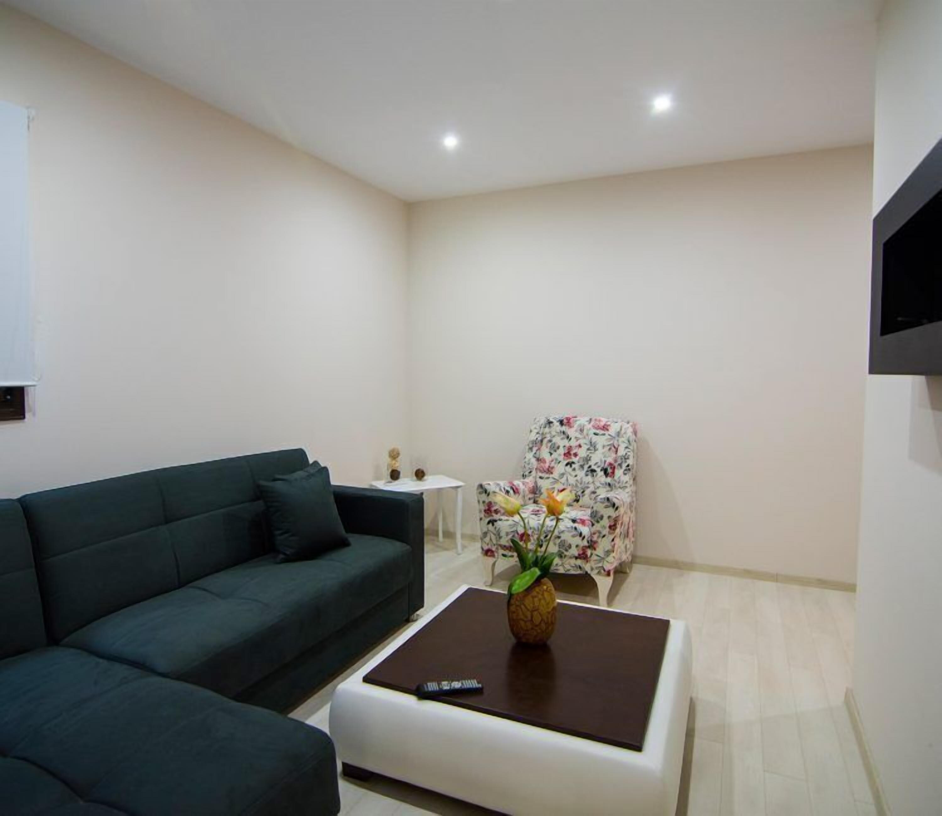 Een moderne woonkamer met een donkergroene bank, een witte salontafel met een afstandsbediening, en een fauteuil met bloemenprint.