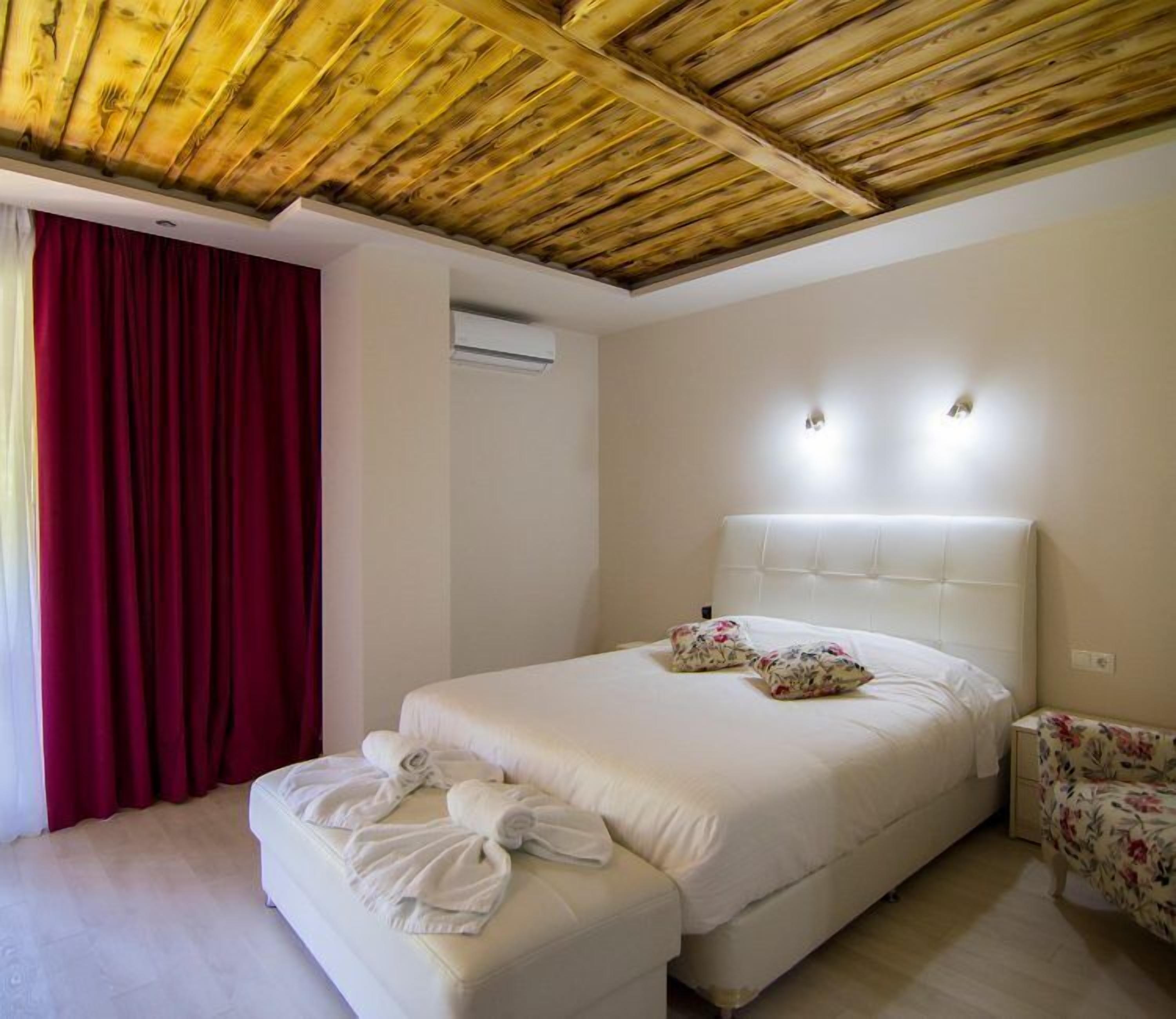 A hotel room with a wooden ceiling, a large bed with white bedding, two wall-mounted lights, and a red curtain.