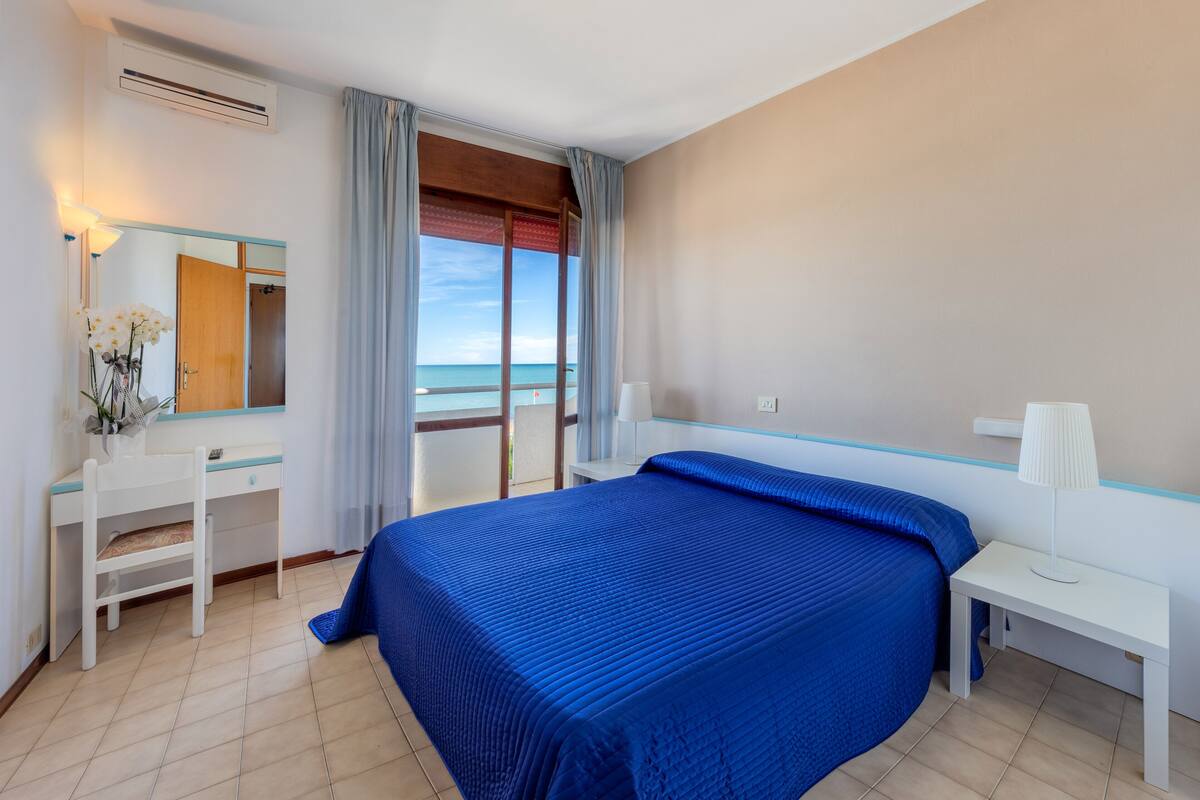 A bedroom with a large bed, a desk, a chair, a mirror, and a view of the sea.