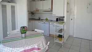 Una cocina compacta con armarios blancos, una mesa de comedor con mantel a rayas y un estante blanco pequeño con una planta.