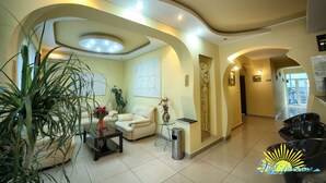 A spacious living area with a sofa, coffee table, and potted plants. There is a trash can and a small table with a vase of flowers. The room has a modern design with curved ceiling lines and recessed lighting. - Vila Diandra (Eforie)