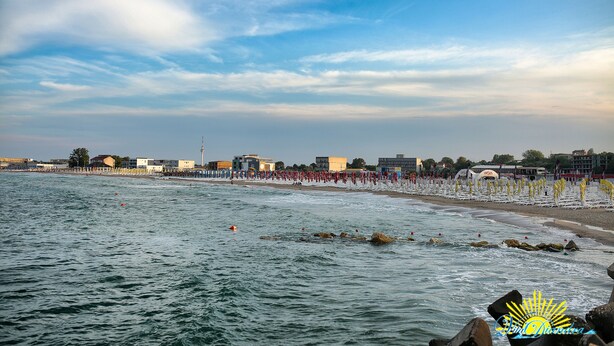 A coastal beach with a row of umbrellas and beach chairs, a rocky shoreline, and buildings in the background. - Vila Diandra (Eforie)
