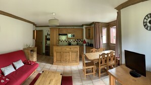 A cozy living room with a red sofa, wooden dining table, and chairs. A kitchen with wooden cabinets and a microwave is visible in the background. - B205 - 58 m2 - 3P - 6 pers (Prapoutel)