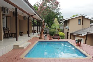A swimming pool with a tiled edge, surrounded by a brick patio. There are steps leading up to a covered area with tables and chairs. - Tzaneen Guest House (Tzaneen)