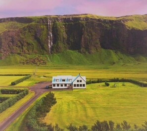 A house with a white exterior and blue roof, surrounded by green fields and a waterfall in the background.
