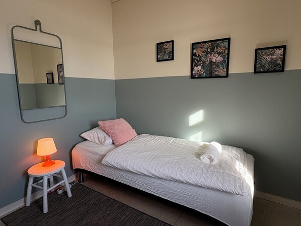 A neatly made bed with white bedding, a small stool with a lamp, and a mirror on the wall.