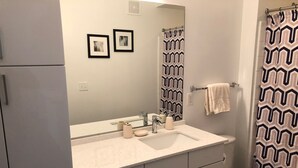 Une salle de bain avec un lavabo blanc, un miroir et des toilettes. Deux tableaux encadrés sont accrochés au mur, ainsi qu’un rideau de douche à motifs géométriques et une serviette.