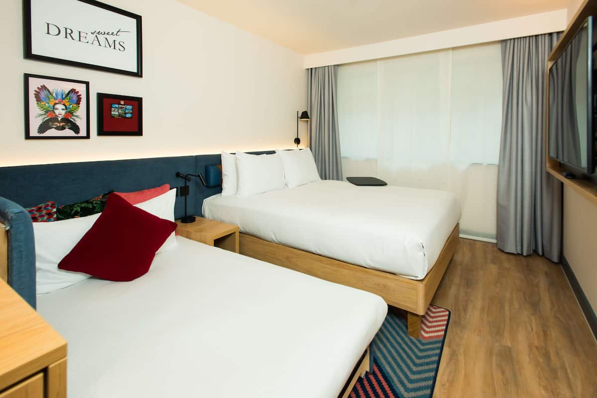 A hotel room with two beds, a large window with curtains, a wall-mounted TV, and framed artwork.