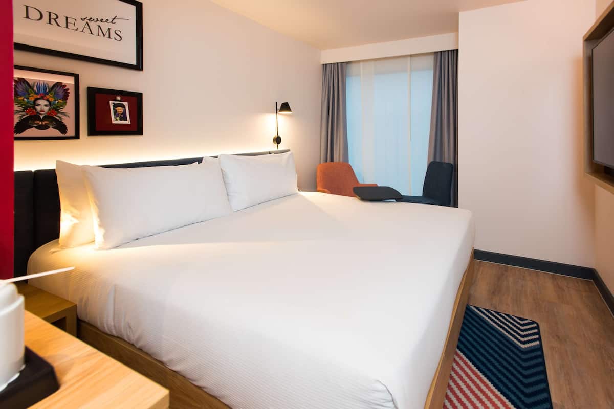 A hotel room with a large bed, a desk with a lamp, a chair, and a TV mounted on the wall.