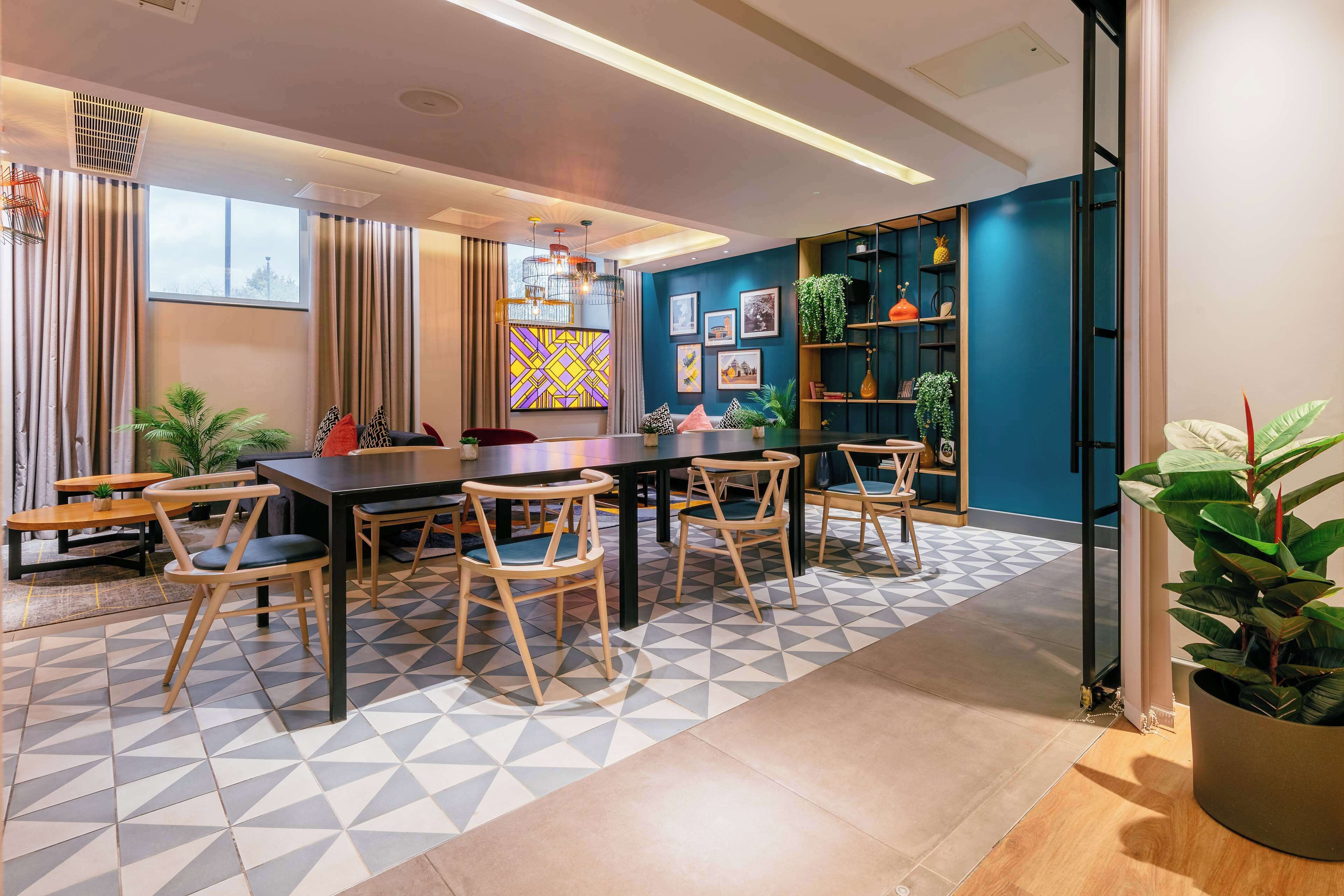 A modern dining area with a black table and wooden chairs, a geometric patterned floor, and a blue accent wall with shelves and plants.
