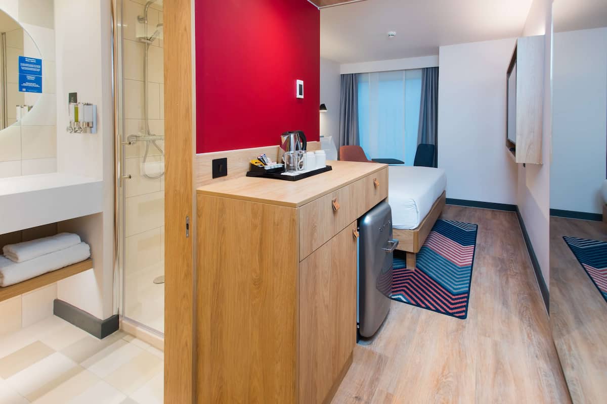 A hotel room with a shower, a bed, a desk with a coffee maker, and a red accent wall.