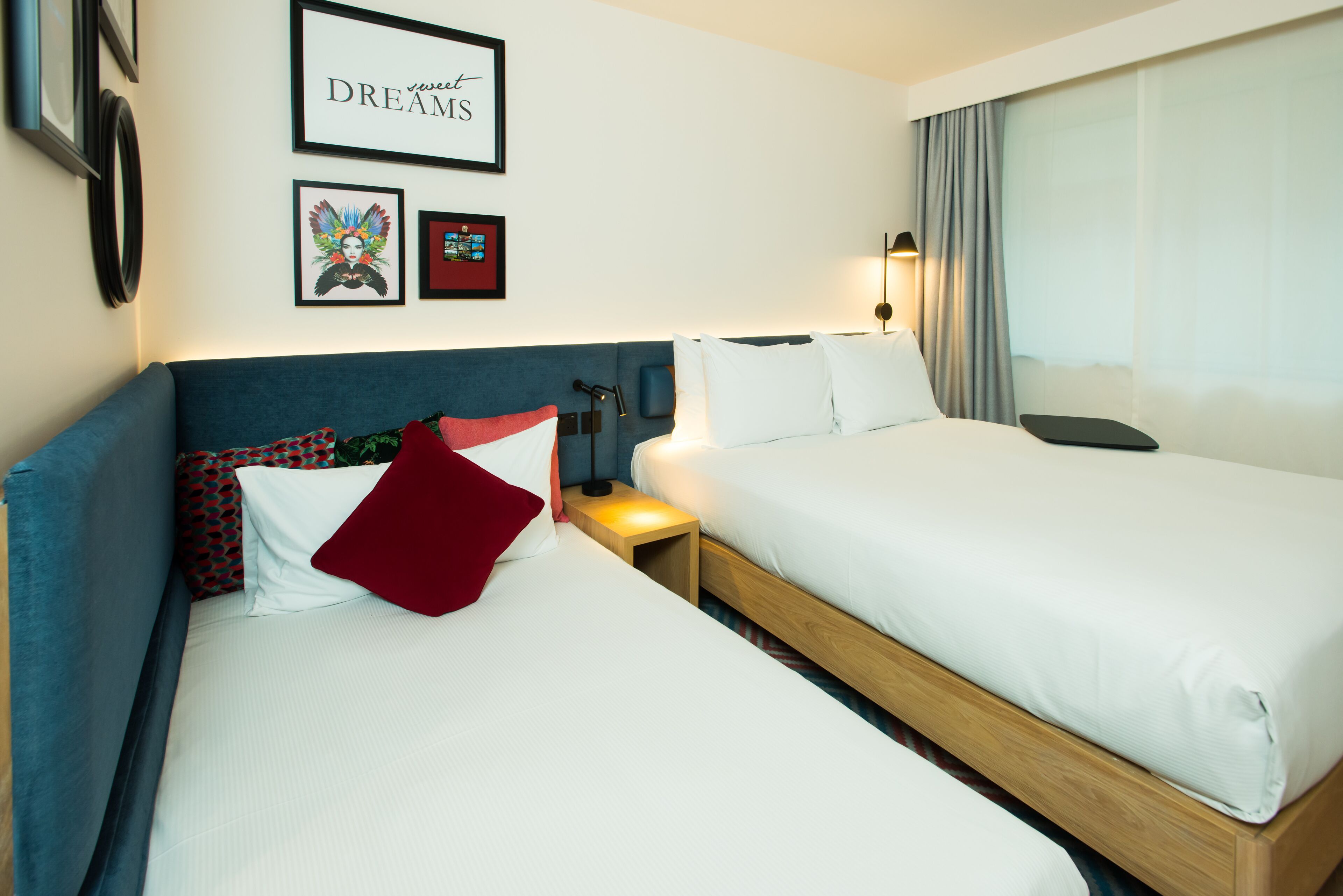 A hotel room with two beds, a blue headboard, a bedside table with a lamp, and framed pictures on the wall.