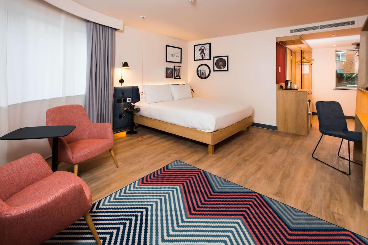 A hotel room with a bed, two red chairs, a desk, a dresser, and a rug.