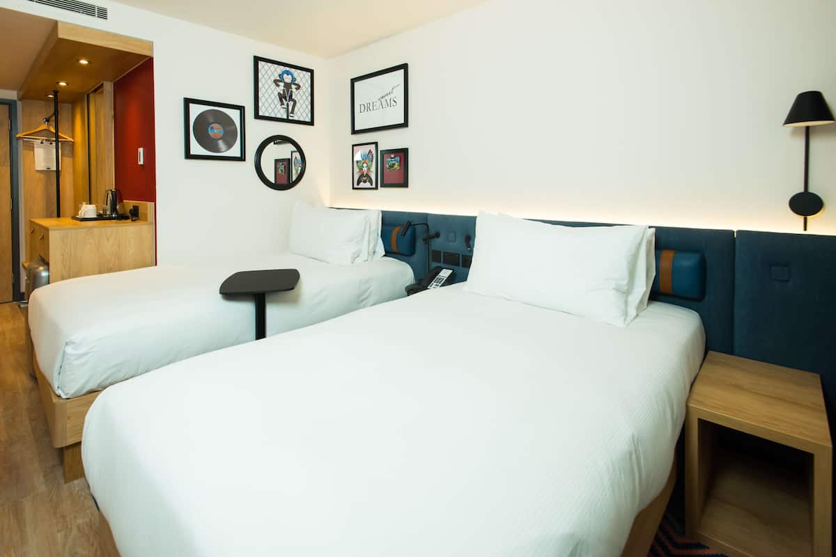 A hotel room with two single beds, a bedside table, a desk, a chair, and a small seating area.