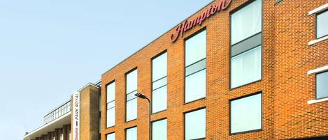 A modern brick building with the Hampton by Hilton logo, a parking lot, and a visible entrance to the Park Royal Hotel.