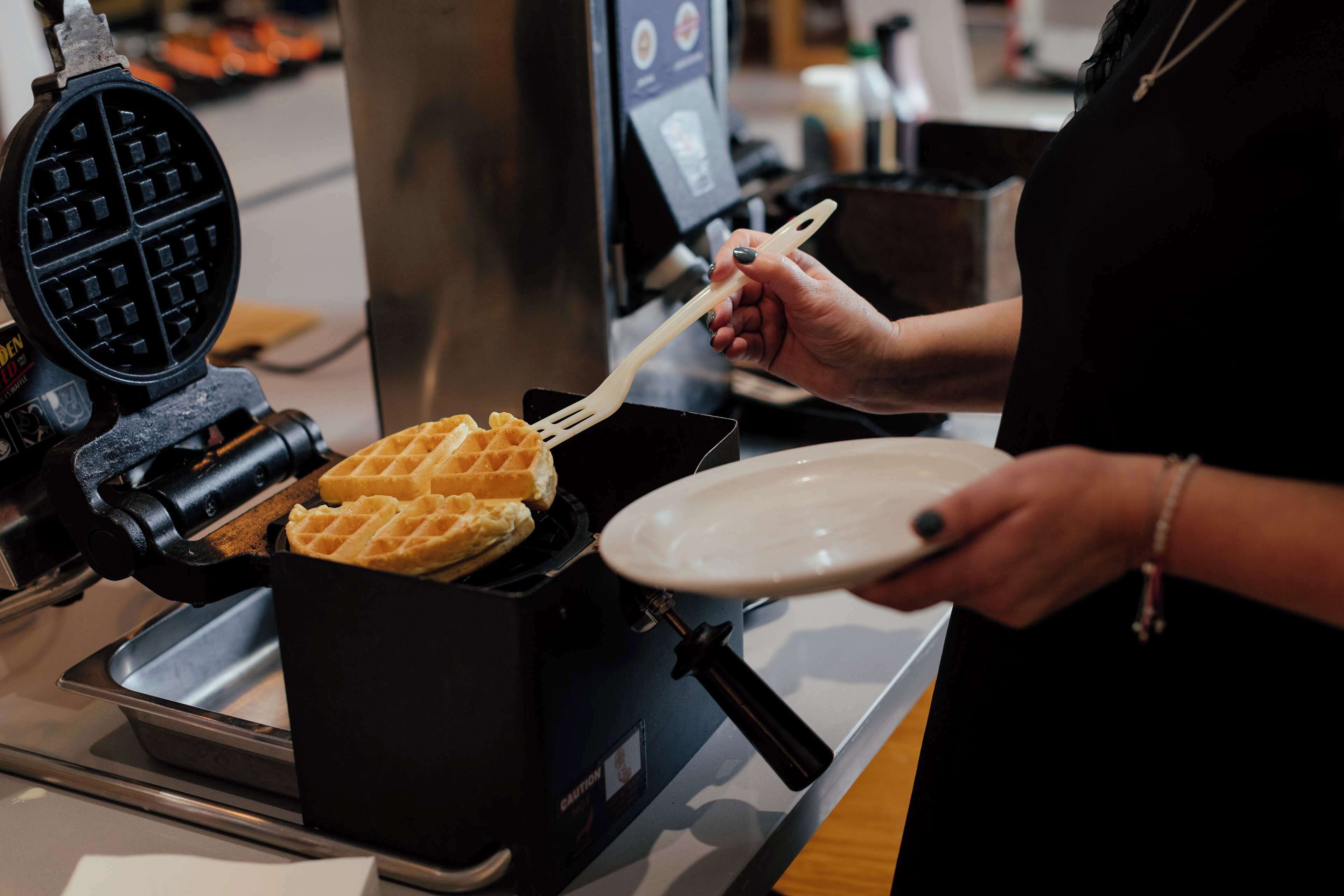 A waffle maker in operation with a person using a spatula to remove a waffle.