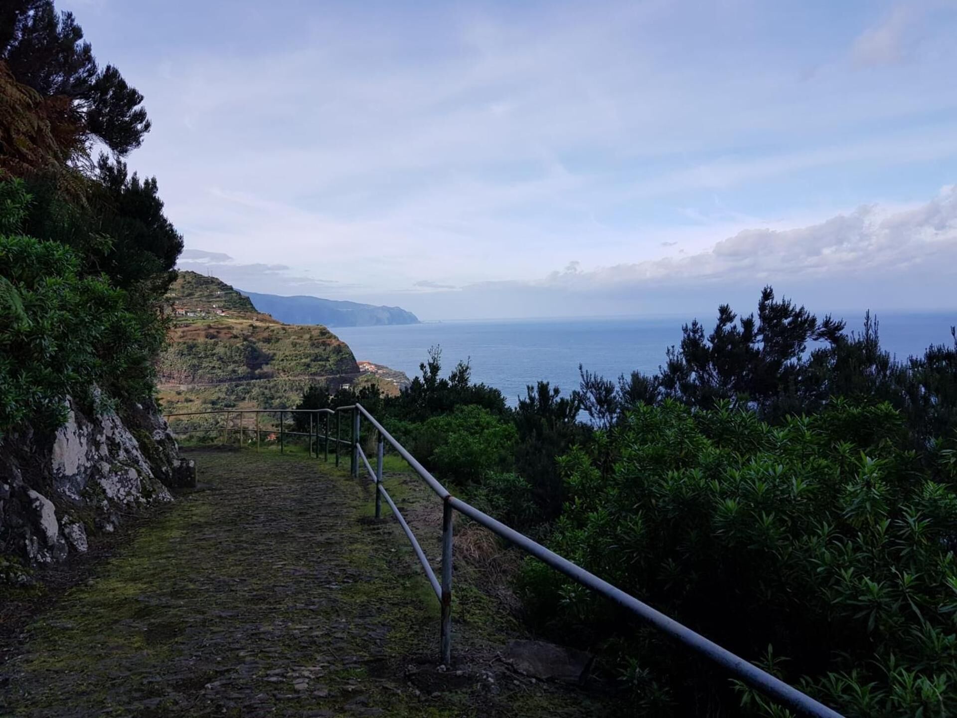 A coastal path with a metal railing, rocky terrain, and a view of the sea and distant hills.