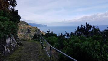 A coastal path with a metal railing, rocky terrain, and a view of the sea and distant hills.