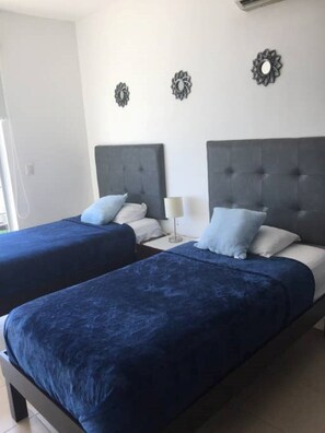 Une chambre avec deux lits, chacun doté d’une tête de lit sombre et d’une literie bleue. Il y a une table de chevet avec une lampe et une décoration murale au-dessus de chaque lit.