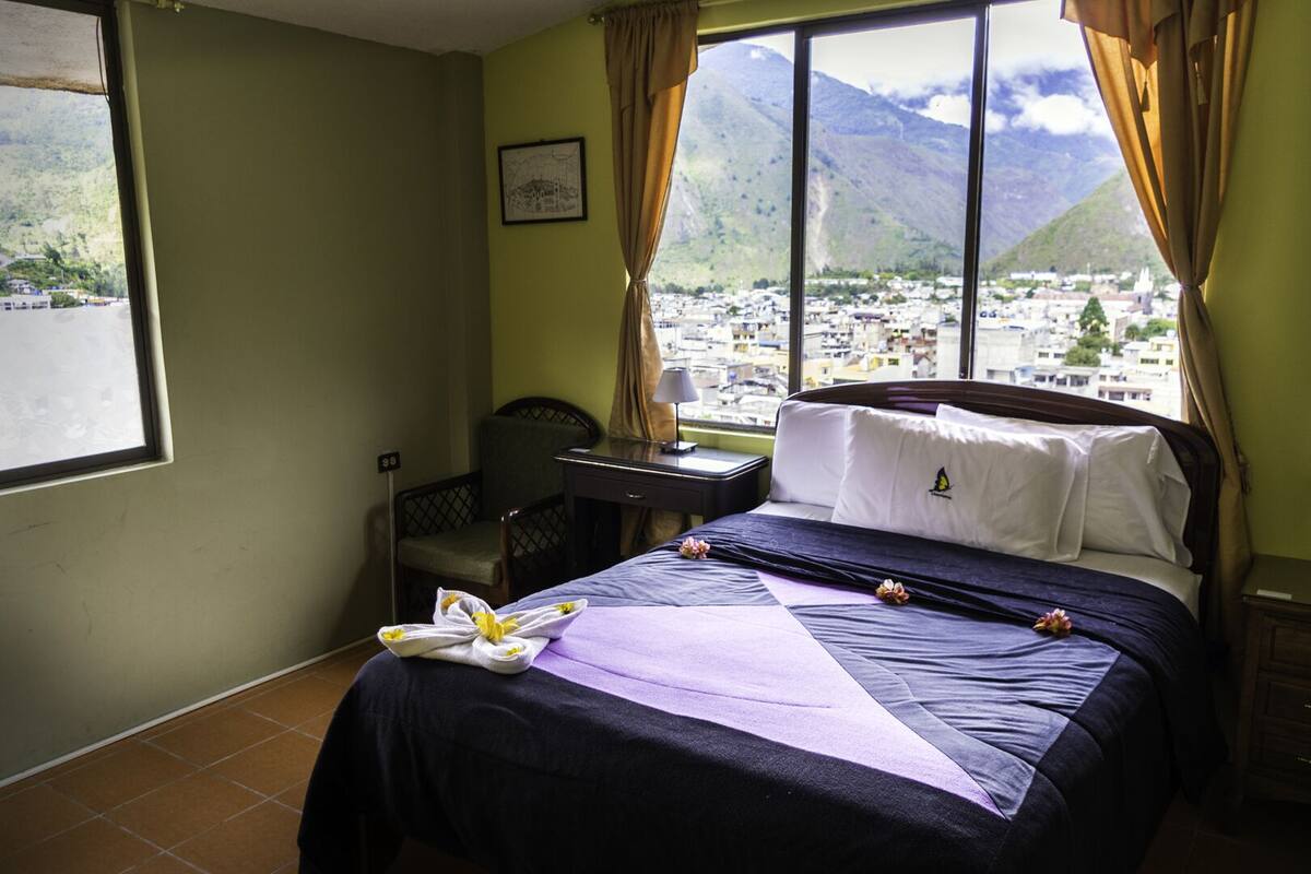 A hotel room with a bed, a chair, a desk, and a large window with a mountain view.