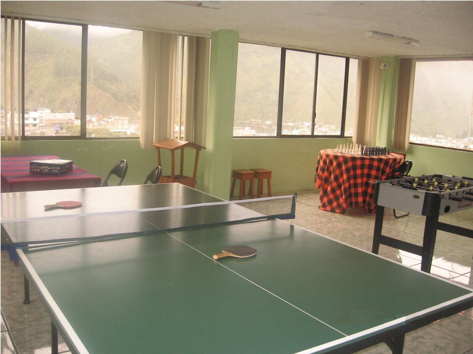 A ping pong table with two paddles, a foosball table, and a table tennis table with a net in a room with large windows.