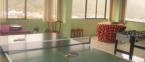 A ping pong table with two paddles, a foosball table, and a table tennis table with a net in a room with large windows.