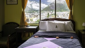 A hotel room with a bed, a desk, a chair, and a window with a view of a city and mountains. - Hosteria Llanovientos (Baños de Agua Santa)