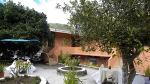 An outdoor patio area with tables and chairs, surrounded by potted plants and a building in the background. - Hosteria Llanovientos (Baños de Agua Santa)