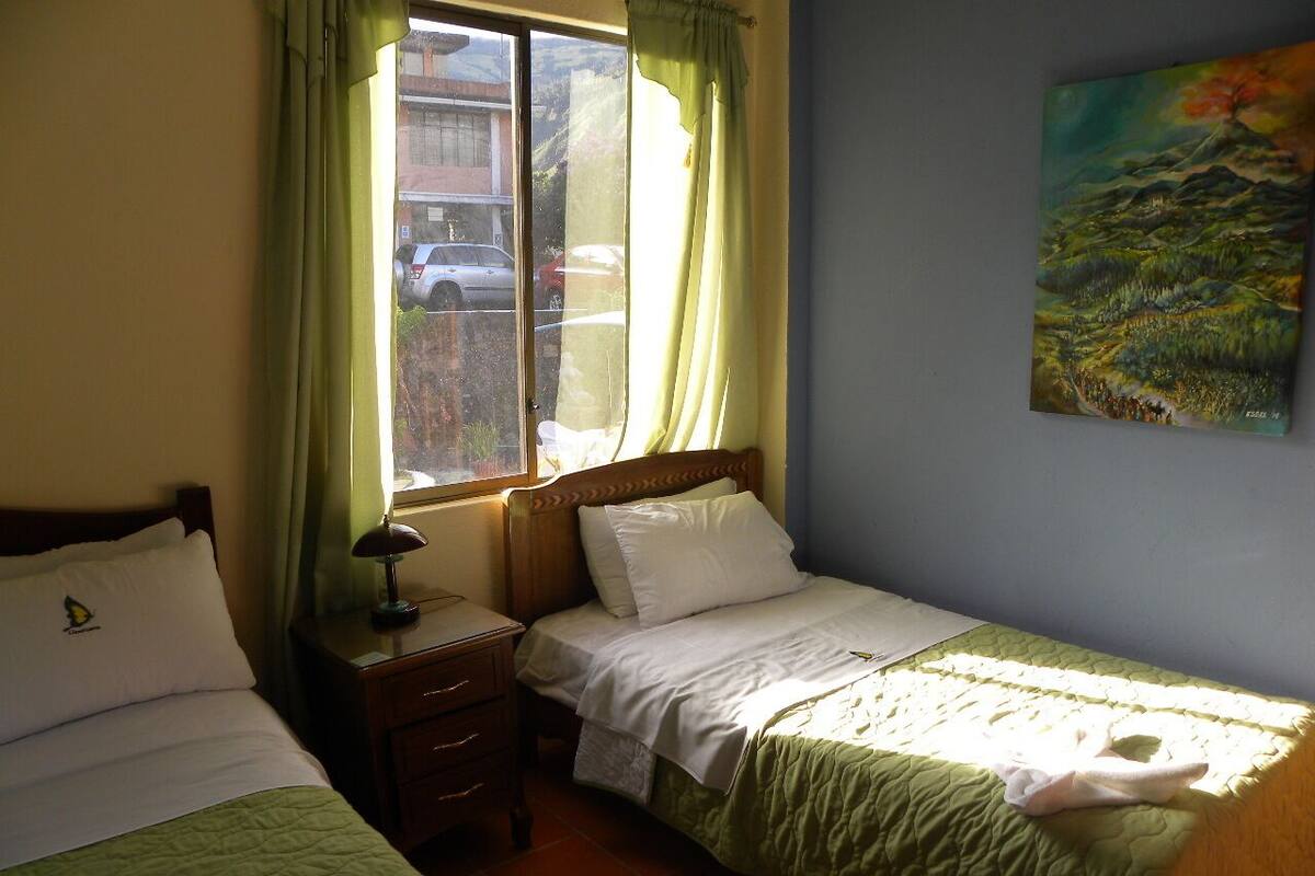 A room with two single beds, a nightstand with a lamp, a window with curtains, and a painting on the wall.