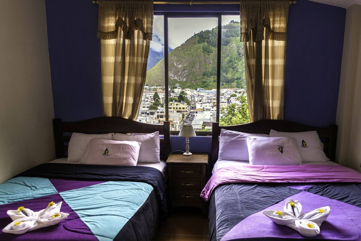A hotel room with two beds, a view of a cityscape and mountains, and a window with curtains.