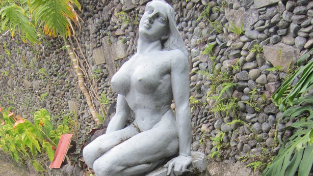 Una estatua de piedra de una figura sentada en un estanque, rodeada de rocas y follaje.