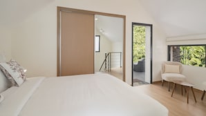 Une chambre moderne avec un grand lit, une armoire en bois, un balcon donnant sur un espace verdoyant et un coin salon comprenant un fauteuil et un canapé.