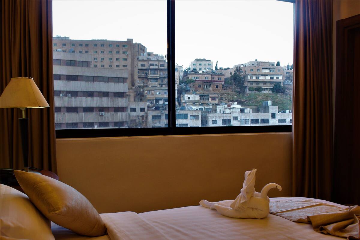 Ein Hotelzimmer mit Bett, Kissen, einer Lampe und Blick auf Stadtgebäude durch ein Fenster.