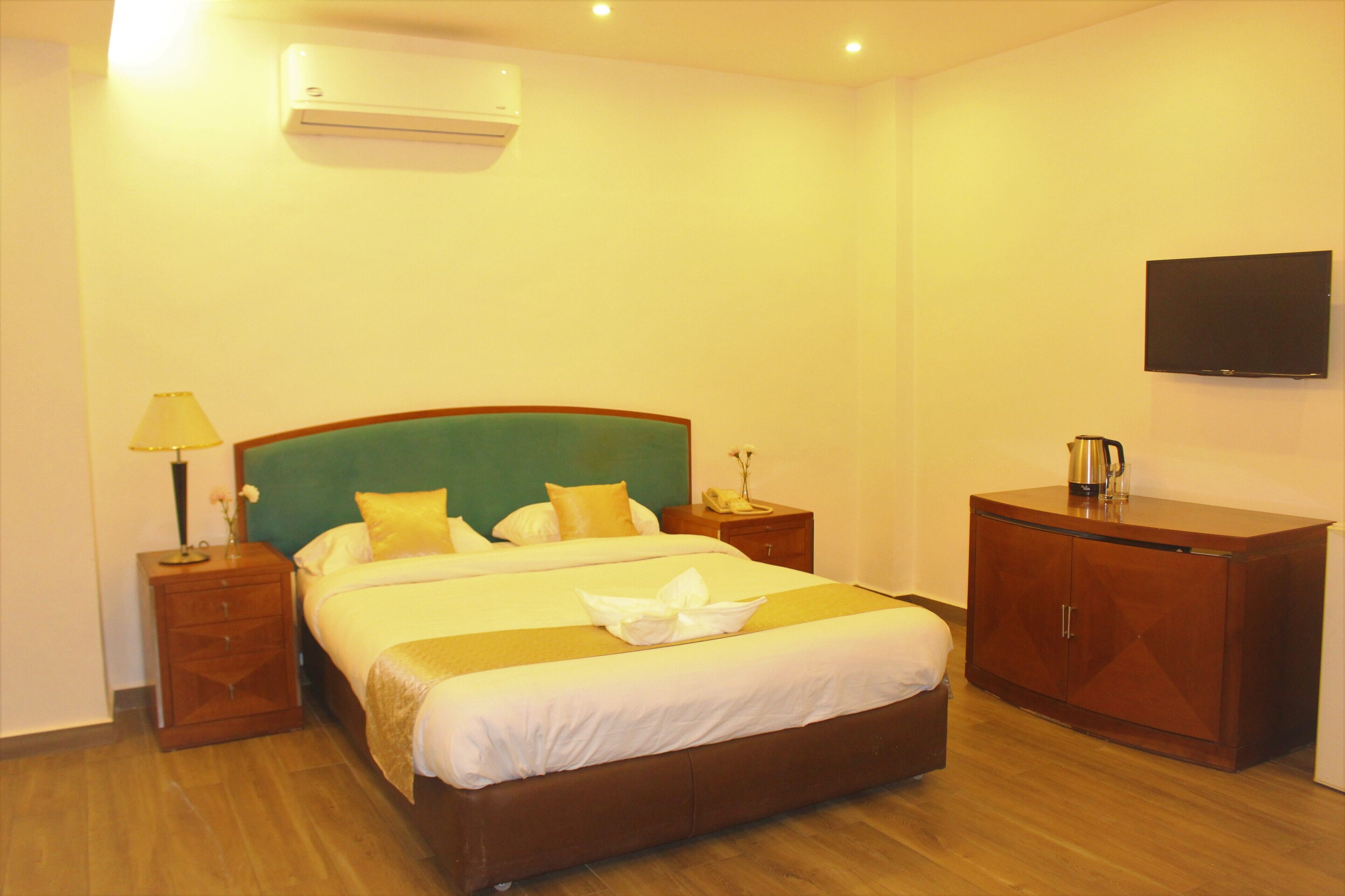 A hotel room with a large bed, a wooden nightstand, a bedside table, a wardrobe, and a flat-screen TV on the wall.