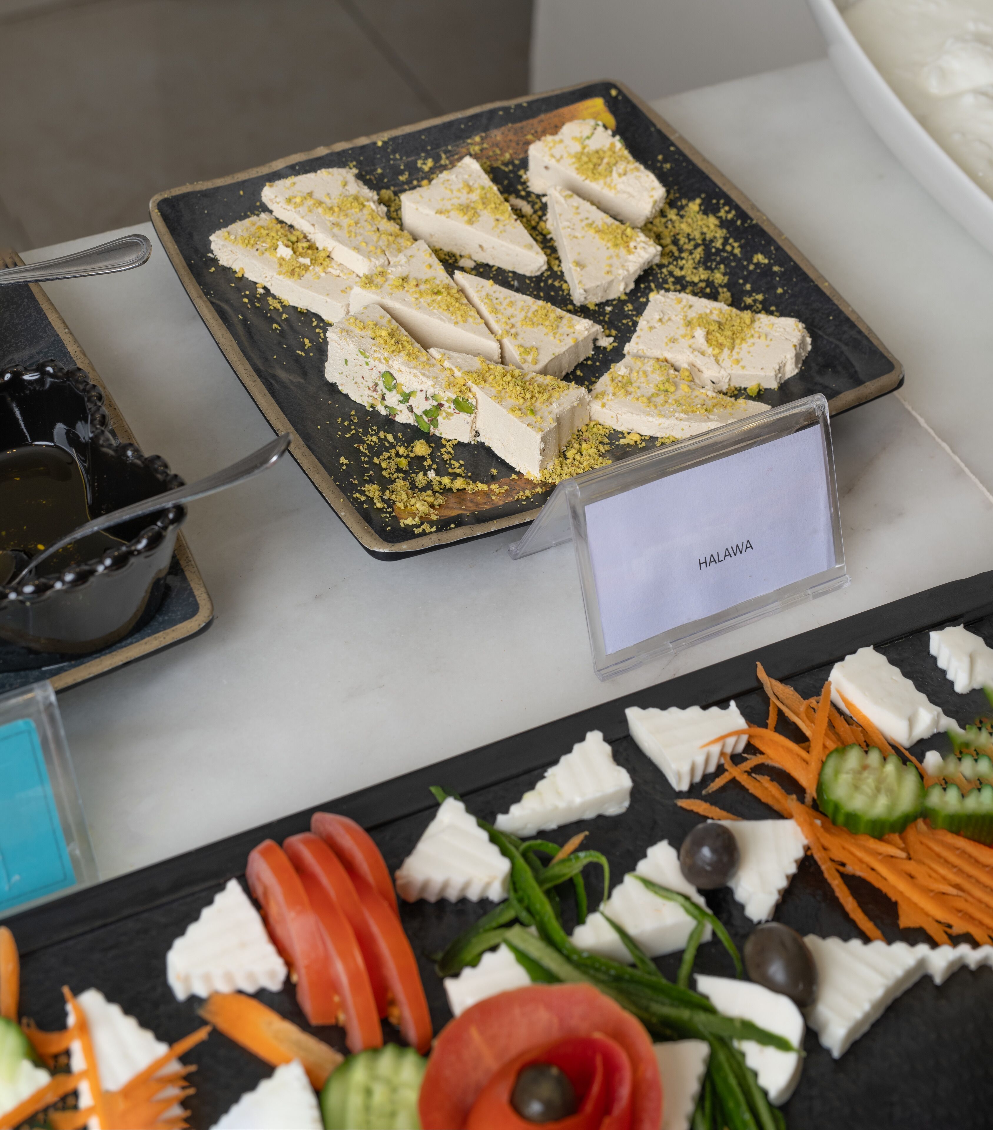 A dessert tray with white cake pieces garnished with green and yellow toppings, labeled "HALAWA," and a variety of fresh vegetables and olives on a black slate.