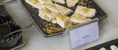 A dessert tray with white cake pieces garnished with green and yellow toppings, labeled "HALAWA," and a variety of fresh vegetables and olives on a black slate.