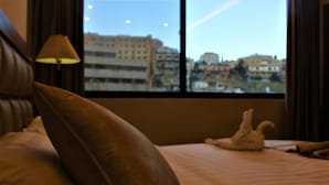 A hotel room with a bed, a lamp, and a view of buildings through the window. - New MerryLand Hotel (Amman)