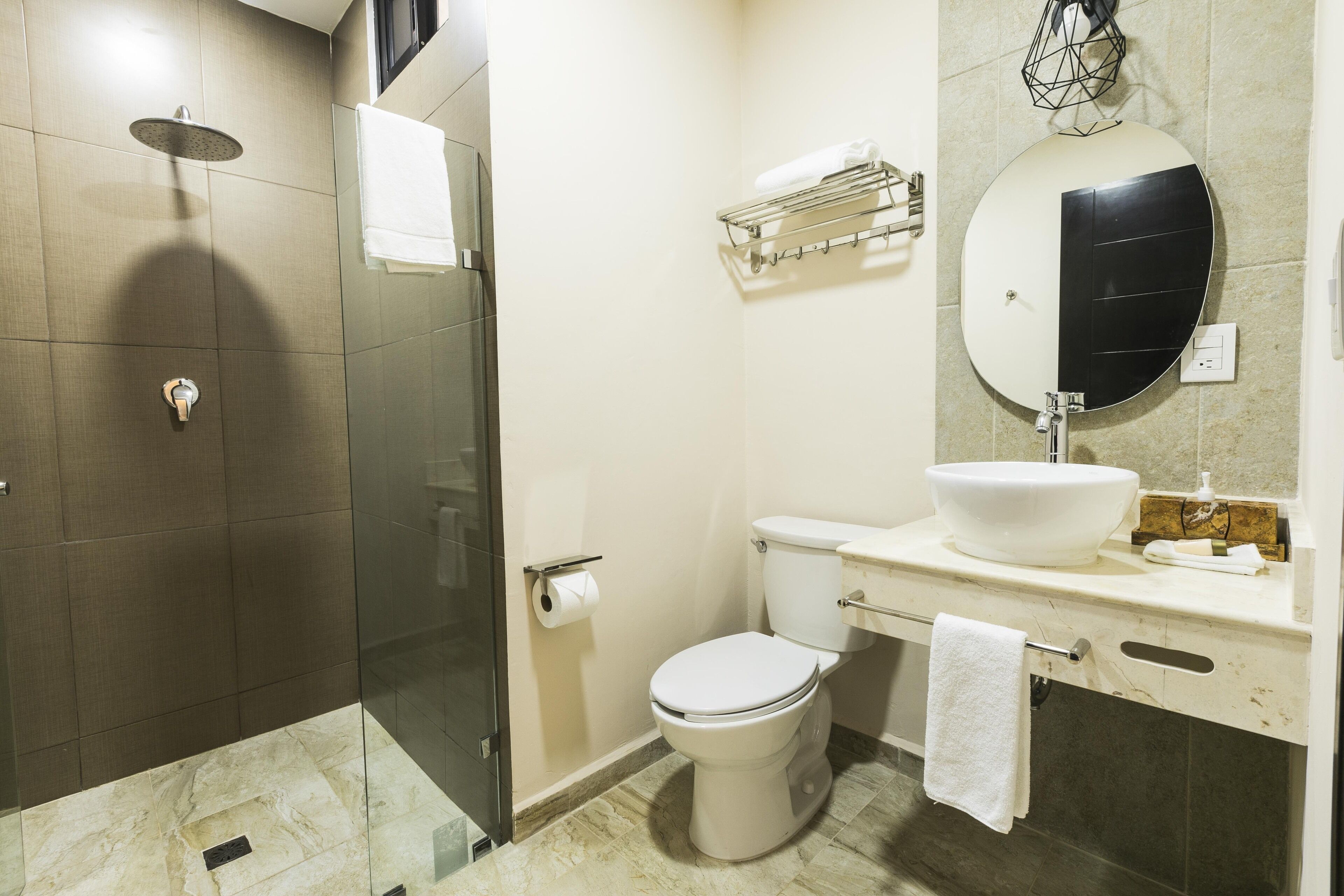A modern bathroom with a glass-enclosed shower, a white toilet, a large round mirror, and a beige countertop with a sink.