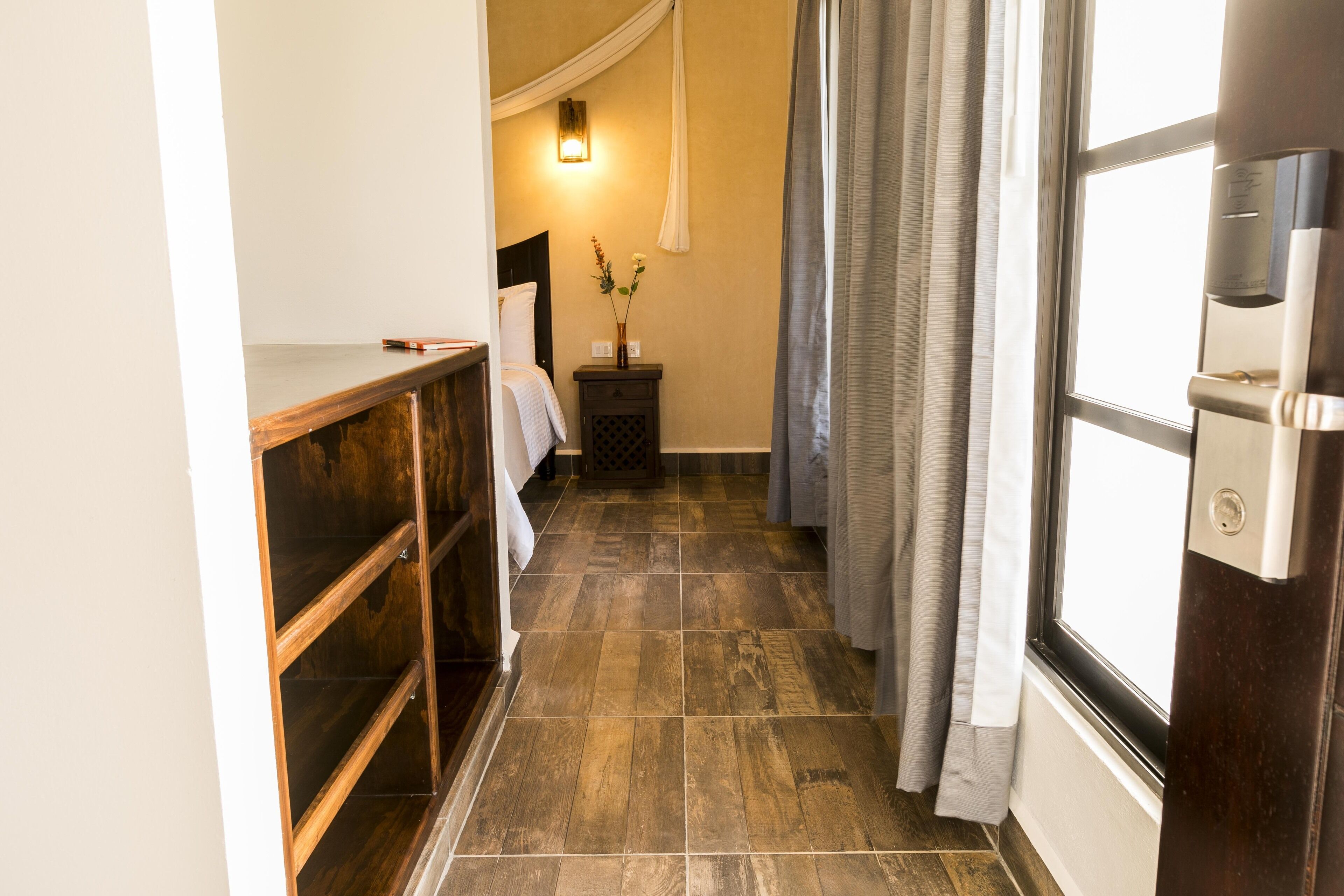 A narrow hallway with a wooden bench, a bedside table with a vase of flowers, a bed with white linens, and a window with curtains.
