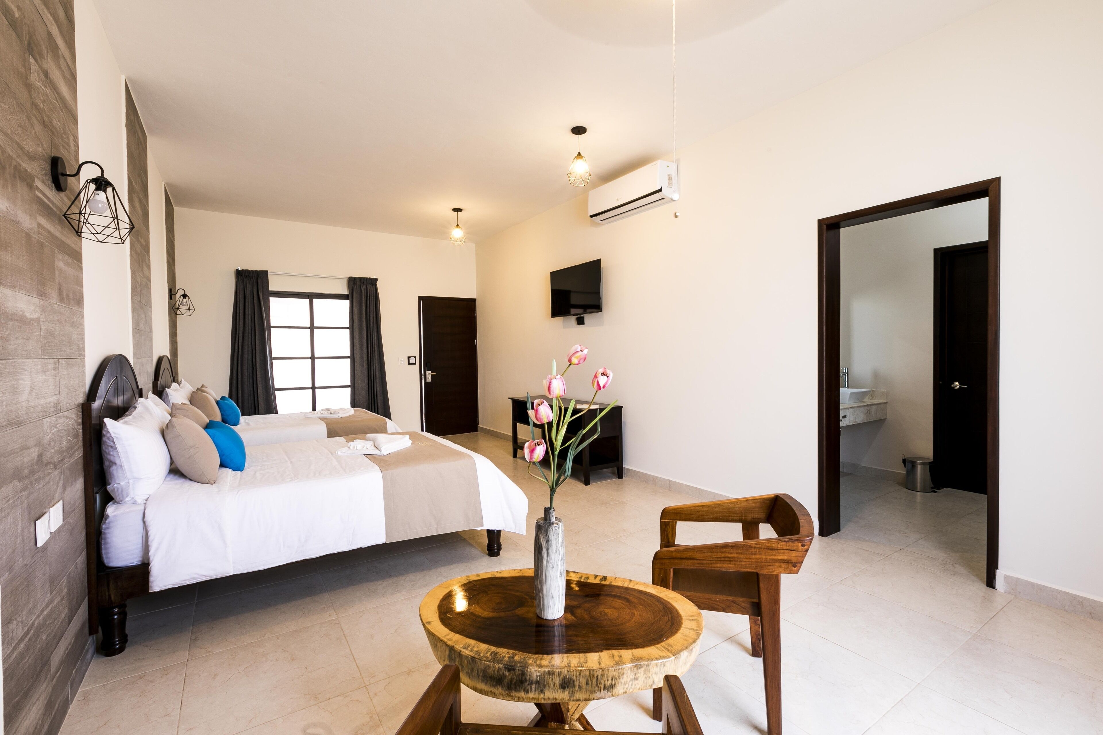 Una habitación de hotel espaciosa con una cama grande, una mesa redonda de madera con un florero y un televisor montado en la pared, además de un equipo de aire acondicionado.