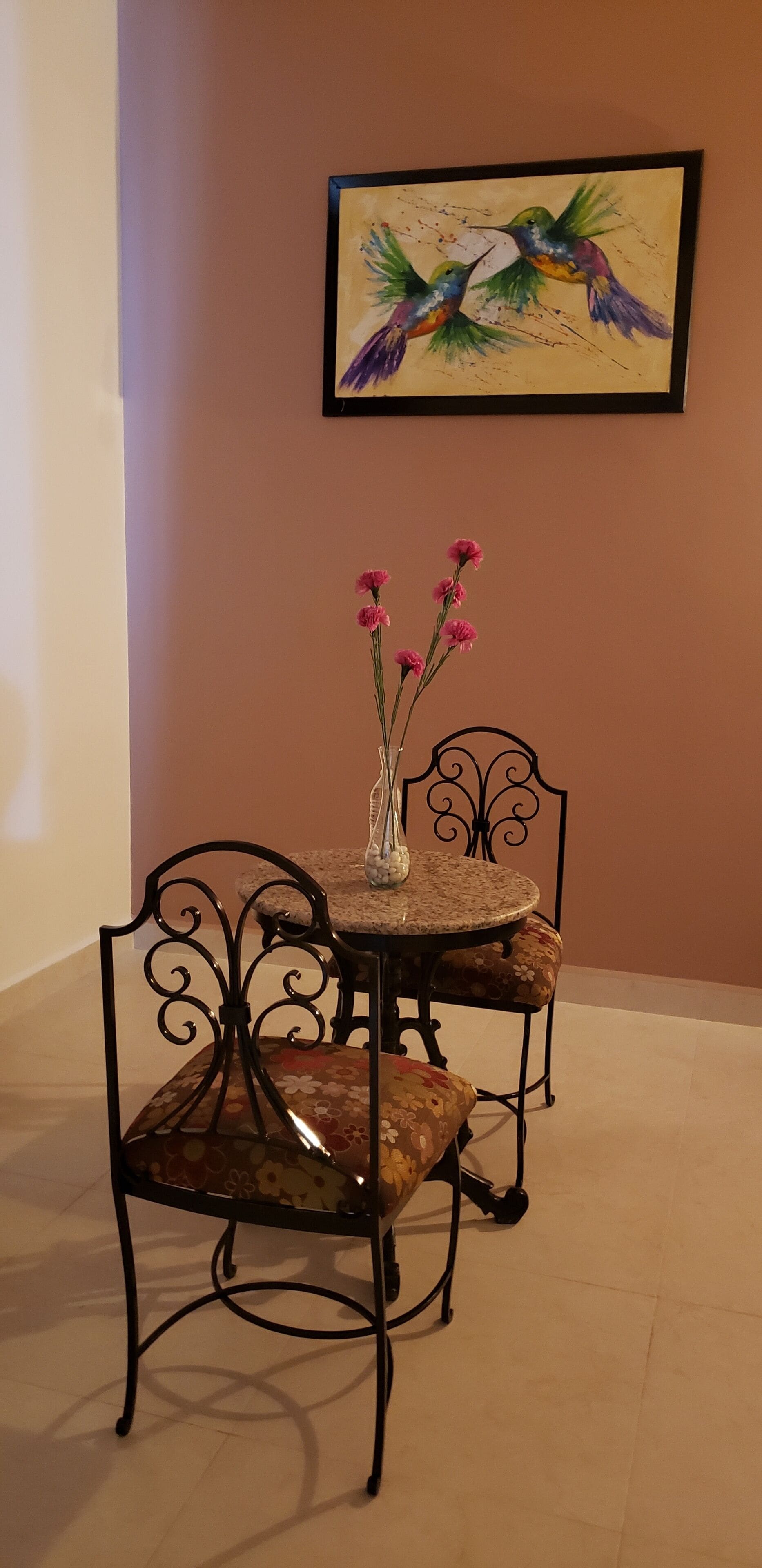 A dining area with a round table and two chairs, a vase with pink flowers, and a framed painting of birds on the wall.