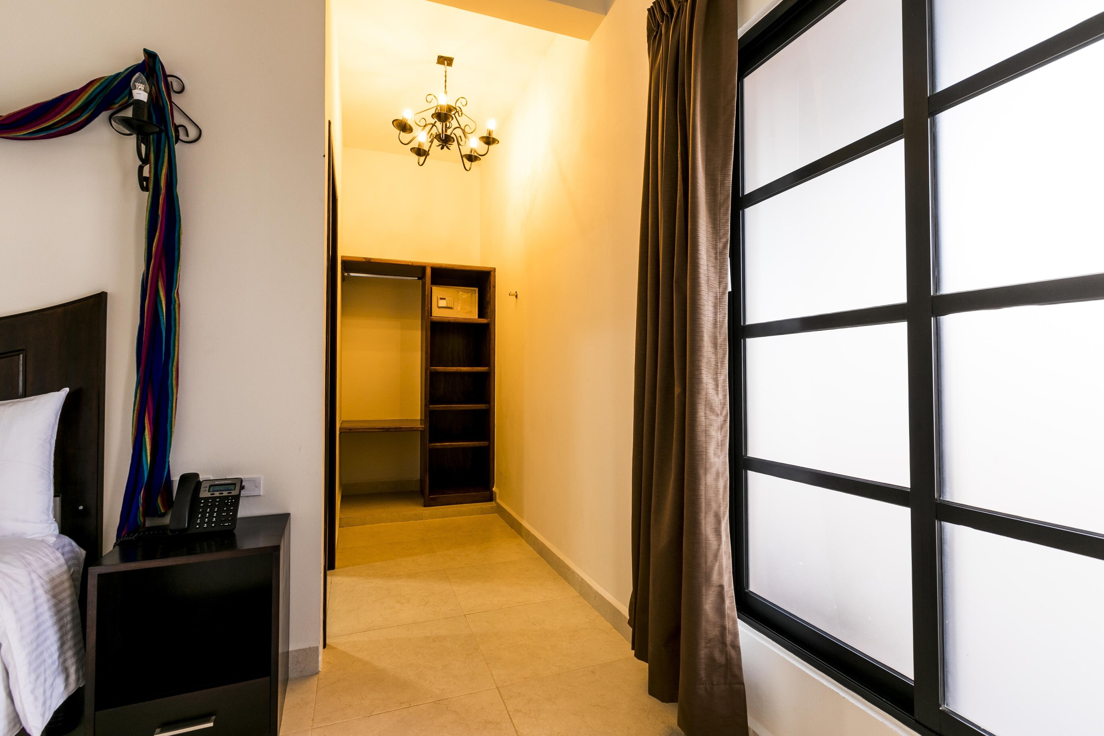 A hotel room with a bed, a bedside telephone, a small desk with a lamp, a wardrobe, and a large window with curtains.