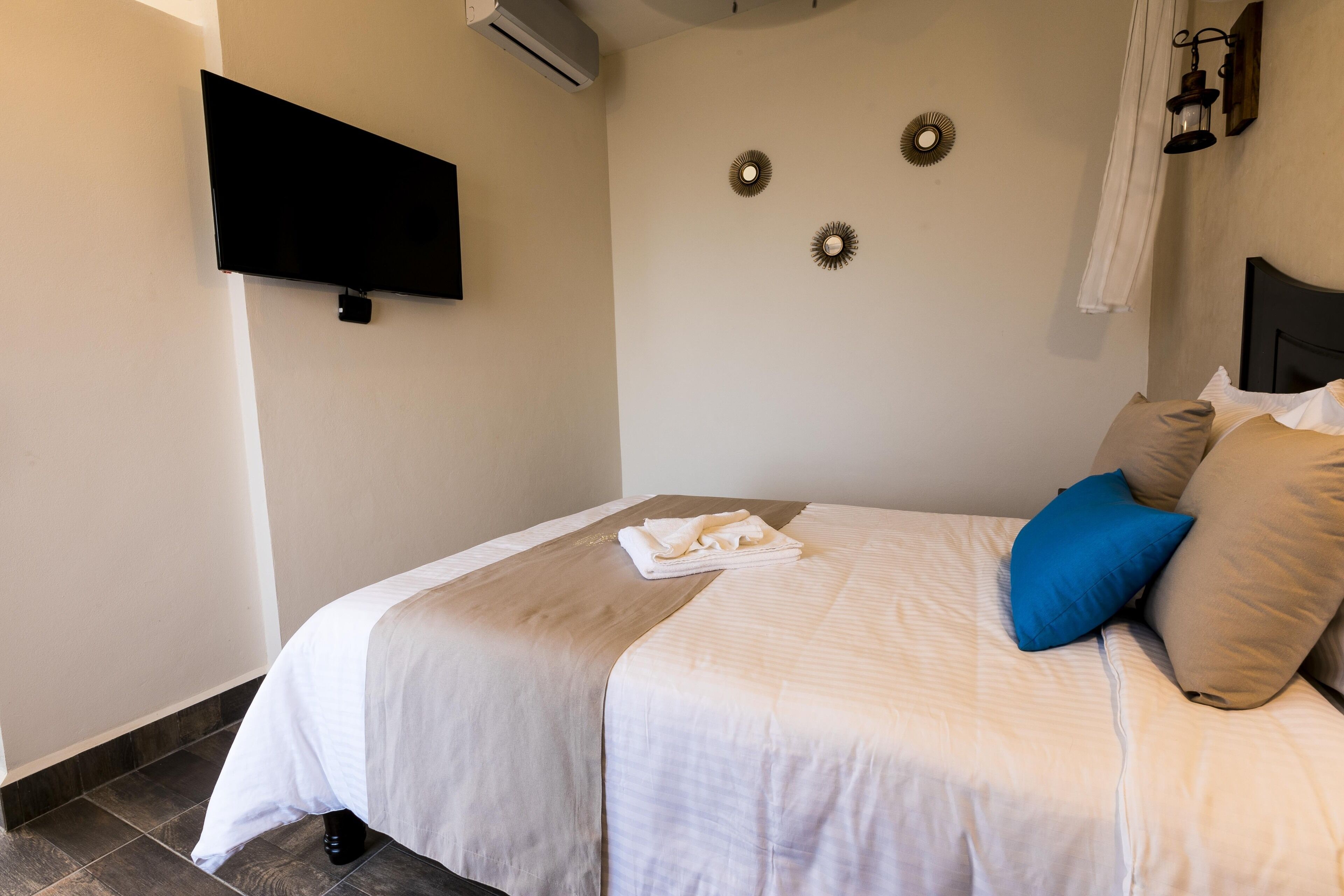 Un lit bien fait, recouvert d’une couverture blanche et beige, avec un oreiller bleu et une serviette blanche pliée. Un téléviseur à écran plat est fixé au mur, au-dessus du lit.