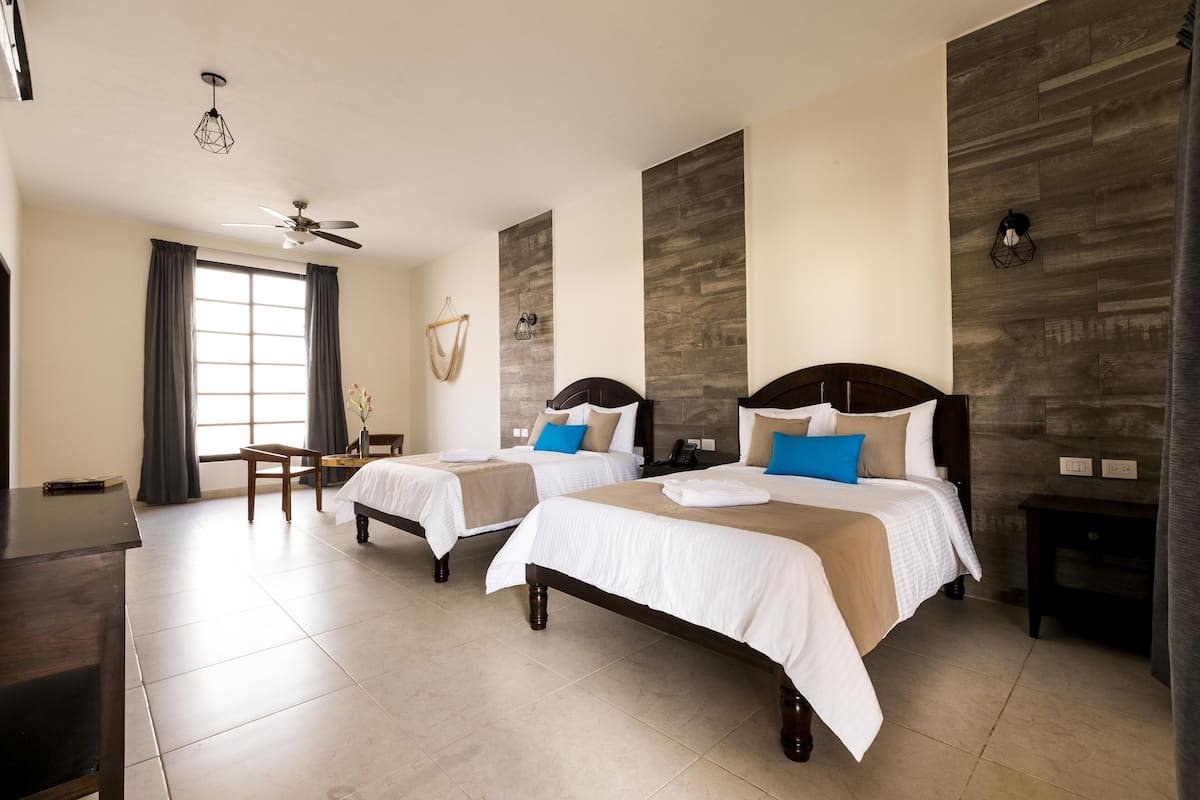 A hotel room with two beds, a desk, a chair, a ceiling fan, and a window with curtains.
