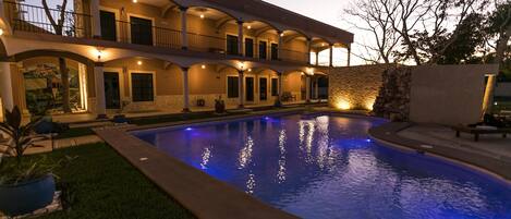Una casa de piscina de varios niveles con una amplia piscina, varios balcones e iluminación exterior.